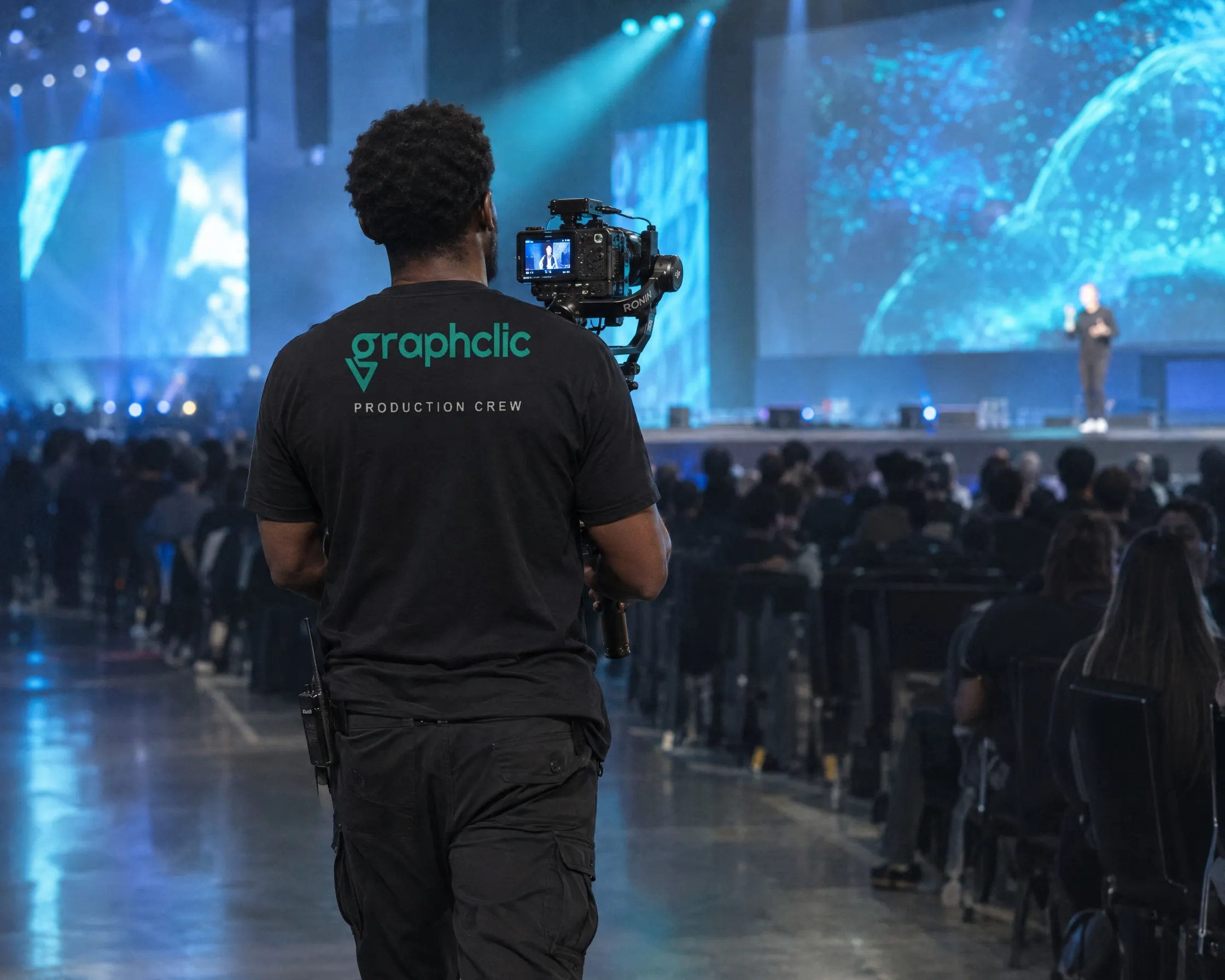 A GraphClic camera operator seen from behind, filming a tech conference stage on a gimbal